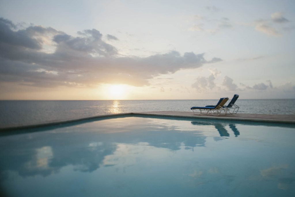Rockhouse Hotel, Negril, Jamaica: Two lounge chairs sit side by side on the edge of an infinity pool overlooking the ocean at sunset, with clouds in the sky and calm water reflecting the soft evening light.
