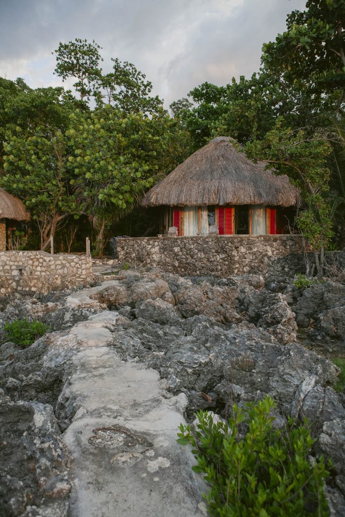 Rockhouse Hotel, Negril, Jamaica: A rustic stone path leads to a round hut with a thatched roof and red window shutters, surrounded by lush green trees and rocky terrain.