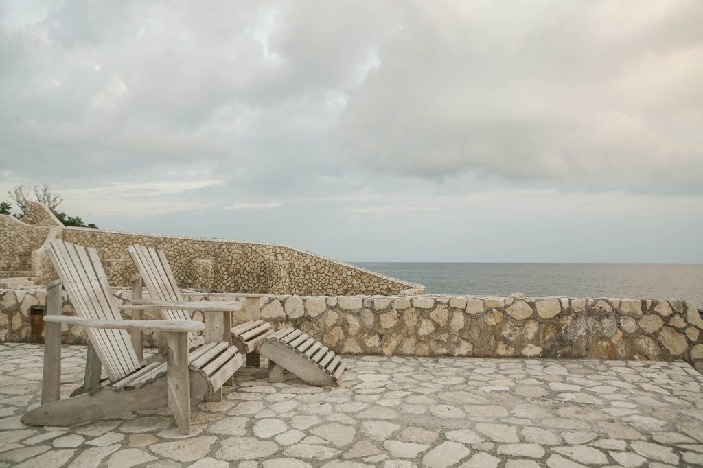 Rockhouse Hotel, Negril, Jamaica: Two wooden lounge chairs sit on a stone patio overlooking a calm ocean, with a stone wall and cloudy sky in the background. The scene appears peaceful and serene.