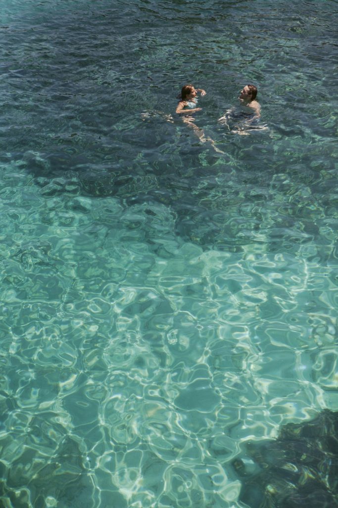 Two people swim in clear, turquoise water under bright sunlight. The shallow areas are light blue, while the deeper areas appear darker. The swimmers are near each other, enjoying the water.