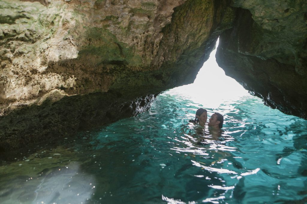 Rockhouse Hotel, Negril, Jamaica: Two people swim together in clear turquoise water under a rocky cave opening, with sunlight streaming through the gap, illuminating the scene.