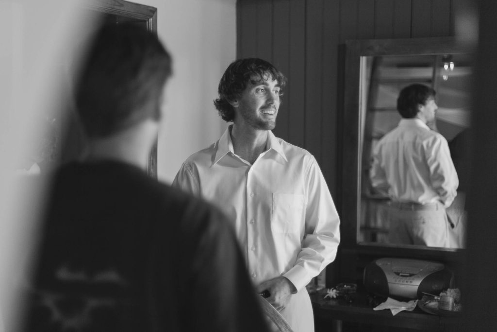 A man in a white dress shirt smiles while standing in front of a mirror. Another person with their back to the camera is in the foreground. The scene appears indoors, with a dresser and stereo visible. The image is in black and white.