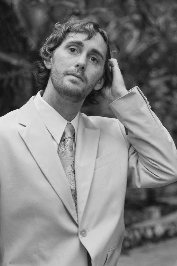 A man in a light-colored suit and patterned tie stands outdoors, looking slightly upward with one hand touching the back of his head. The background is blurred with foliage. The photo is in black and white.