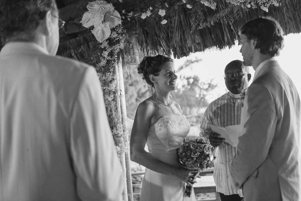 A bride and groom stand under a floral arch during their wedding ceremony. The bride holds a bouquet and smiles at the groom, who is reading from a paper. An officiant and another man stand nearby. The setting is outdoors.