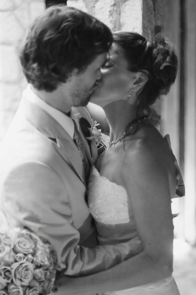 A bride and groom share a kiss, embracing closely. The bride holds a bouquet of roses and wears a strapless dress, while the groom is in a light-colored suit. The image is in black and white.