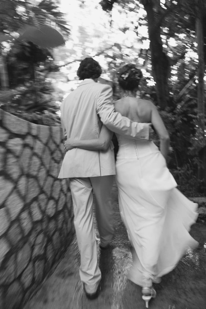 A couple, dressed in wedding attire, walk arm in arm along a stone path surrounded by trees. The image is black and white, with a soft, slightly blurred effect suggesting movement and intimacy.