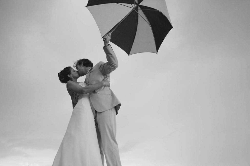A bride and groom kiss passionately as they embrace, holding a large umbrella above them. The sky is overcast, and the image is in black and white, creating a romantic and timeless scene.