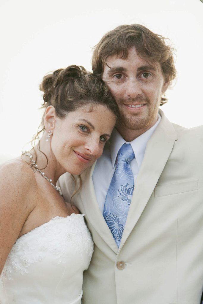 Rockhouse Hotel, Negril, Jamaica: A smiling bride in a strapless white dress leans affectionately into a groom in a light suit and blue patterned tie, both posing closely together against a bright background.