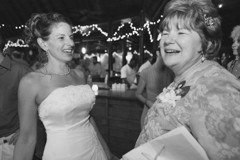 A bride in a strapless wedding dress smiles and talks with an older woman holding papers at a lively indoor reception, with string lights and guests in the background.