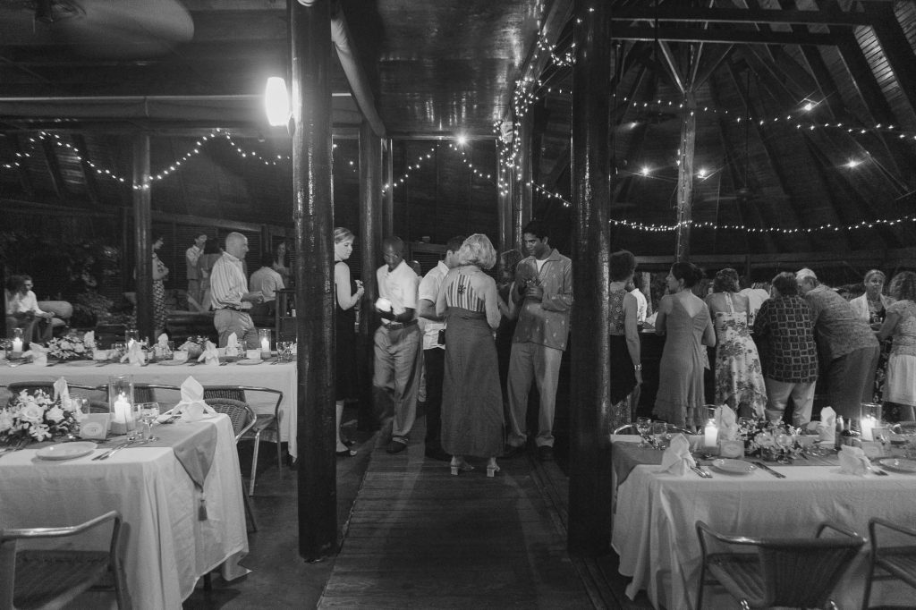 Black and white photo of a group of people socializing in a decorated indoor venue with string lights. Banquet tables with flowers, candles, and napkins are set up on each side of the space.
