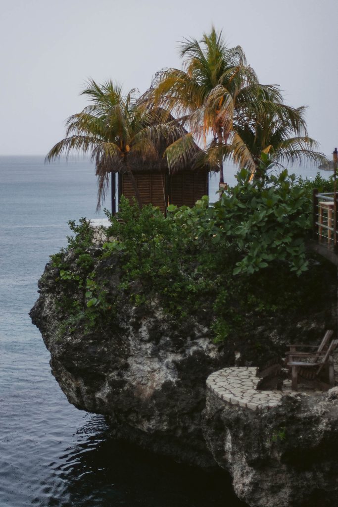 Rockhouse Hotel, Negril, Jamaica: A small wooden hut sits among palm trees on a rocky cliff overlooking a calm ocean, with lush green foliage and a stone patio with wooden chairs nearby.
