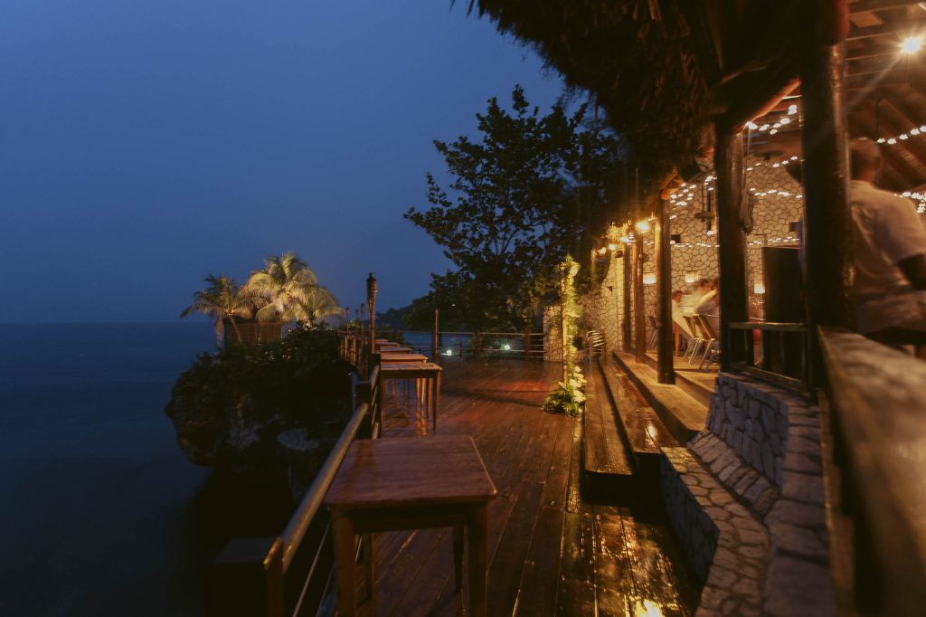 Rockhouse Hotel, Negril, Jamaica: Outdoor dining area on a wooden deck overlooking the ocean at dusk, with string lights and palm trees, creating a warm, inviting tropical ambiance.