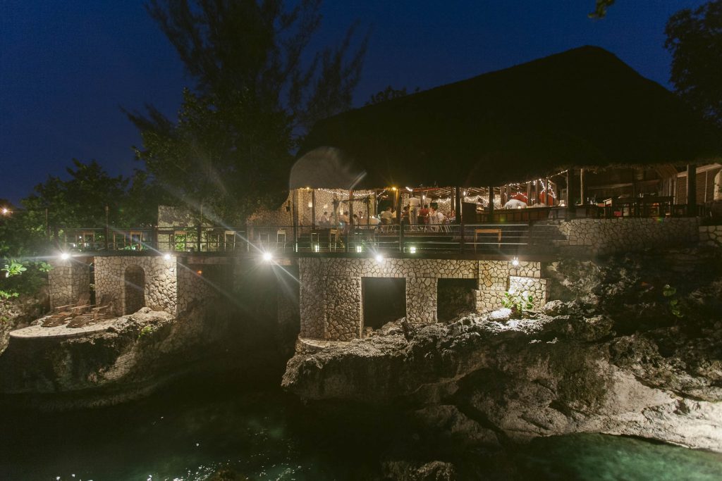 Rockhouse Hotel, Negril, Jamaica: A stone building with a thatched roof sits above rocky cliffs and water, warmly lit at night. A wedding reception is visible inside, and lights illuminate the exterior, creating a cozy, inviting atmosphere surrounded by trees.