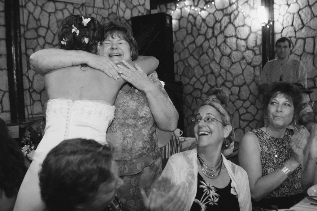 A bride in a strapless gown hugs an older woman who is smiling joyfully. Other guests sitting at a table nearby clap and laugh, enjoying the celebration in a warmly lit, stone-walled venue.
