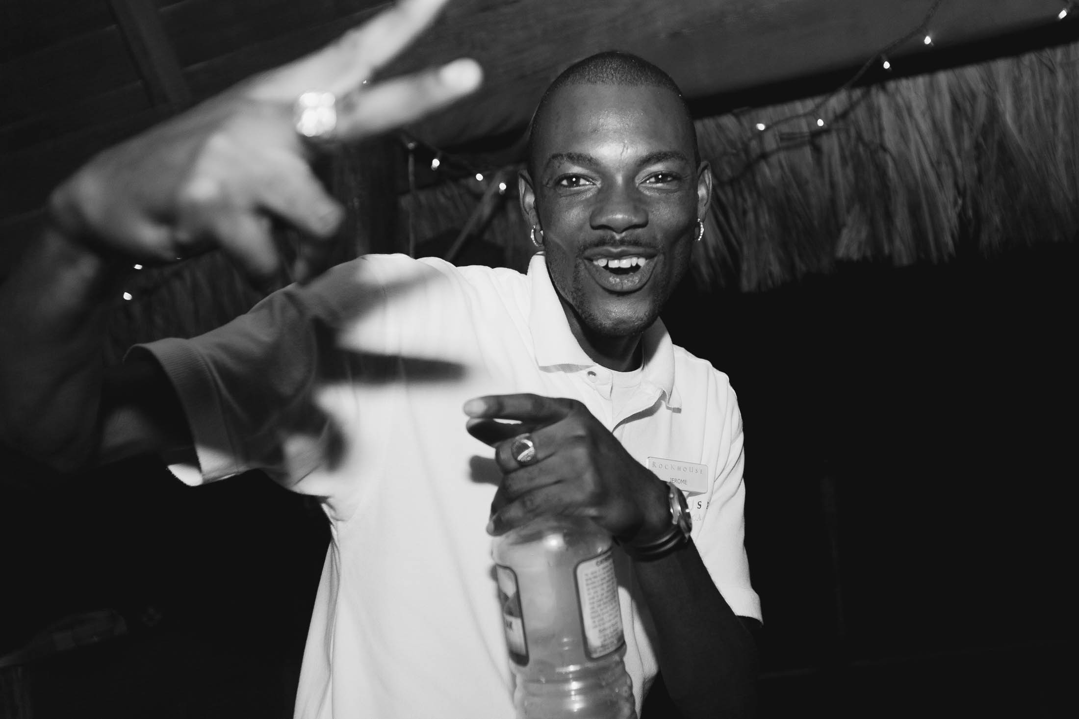 A smiling man in a white shirt holds a bottle and gestures a peace sign with his hand. Strings of small lights hang in the background, and the image is in black and white.