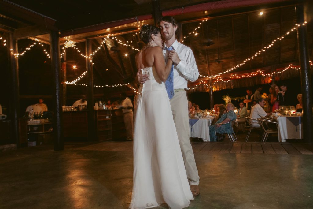 A bride and groom share a slow dance under string lights at a warmly lit indoor wedding reception, with guests seated at decorated tables in the background.