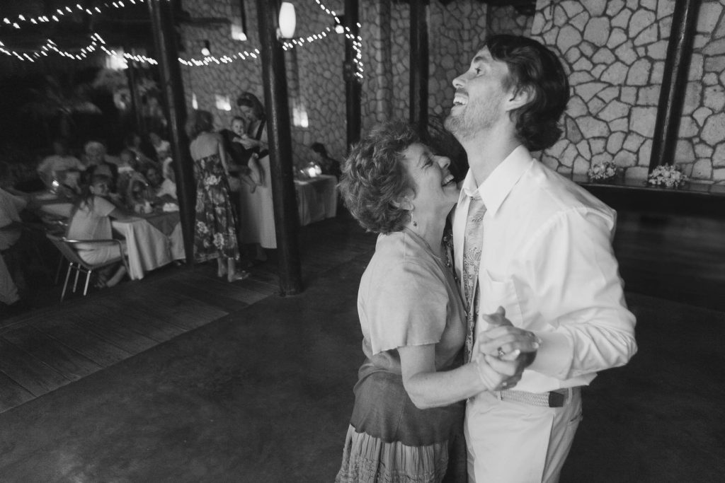 A joyful older woman and a man in a tie dance together and laugh at a festive indoor event, while guests sit at decorated tables in the softly lit background.