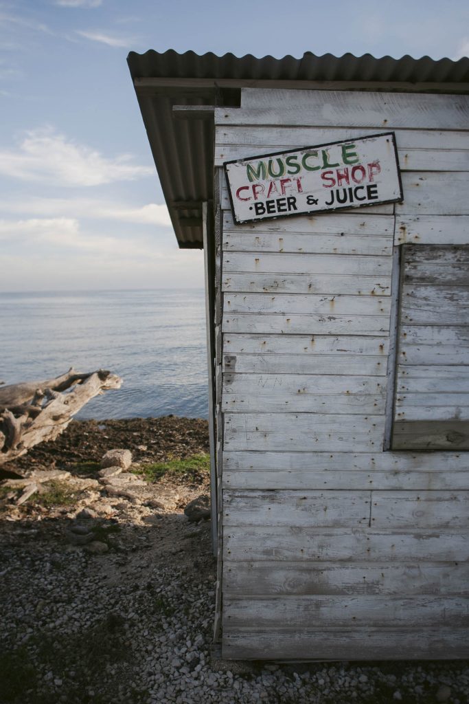 A weathered wooden shack by the sea has a hand-painted sign reading “MUSCLE CRAFT SHOP BEER & JUICE.” Rocky shore and driftwood are visible in the background under a partly cloudy sky.