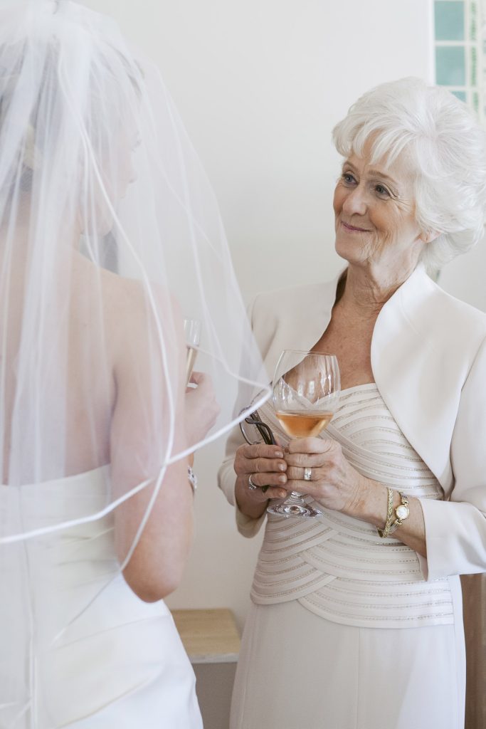 An older woman in an elegant light outfit smiles warmly at a bride in a wedding dress and veil. Both hold glasses of rosé wine and appear to be sharing a pleasant conversation in a bright setting.