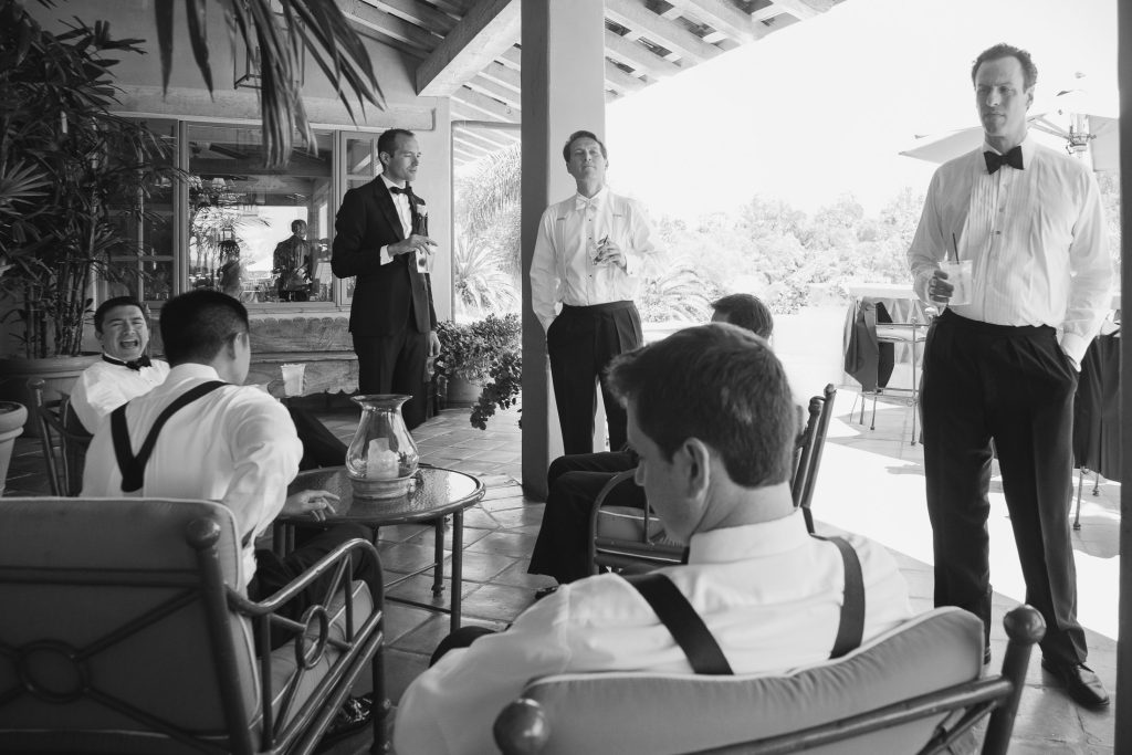 A group of men dressed formally in white shirts and black suspenders or bow ties relax on a patio. Some are sitting and some are standing, holding drinks, having a conversation in a casual, outdoor setting.