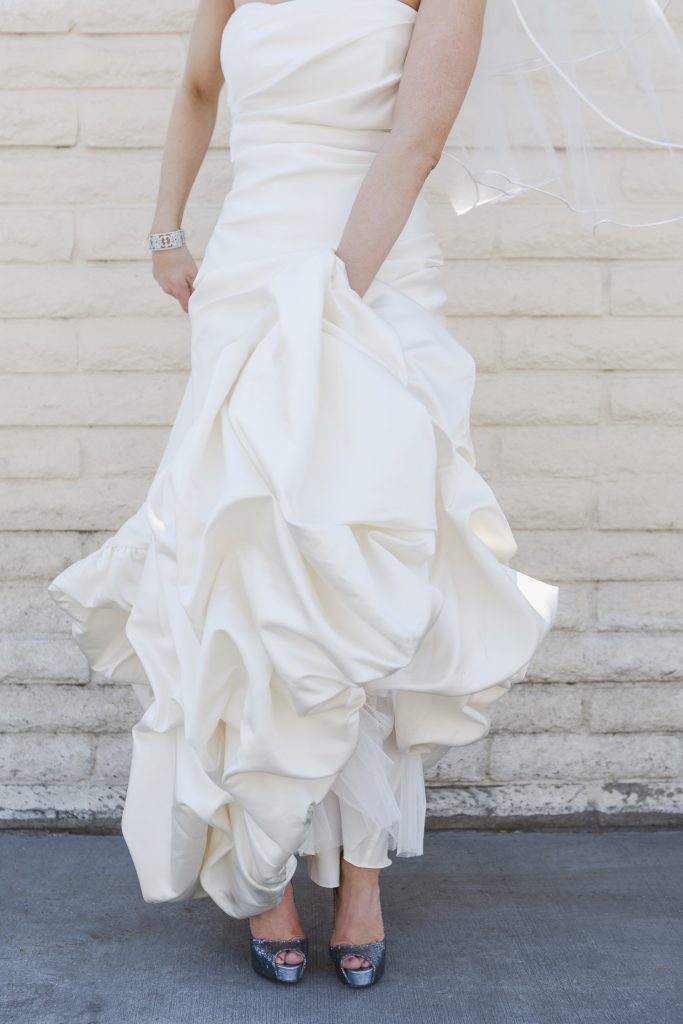 A bride in a strapless white wedding dress lifts her gown slightly, revealing sparkly high heels. She stands in front of a light-colored brick wall, with her veil and bracelet visible. Her face is not shown.