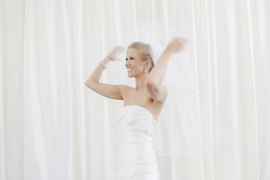A smiling bride in a strapless white wedding dress adjusts her veil against a softly lit white curtain background.