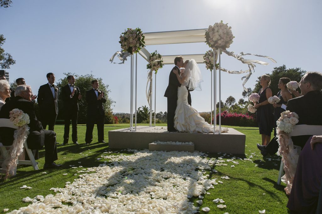 A bride and groom share a kiss under a floral wedding arch outdoors, surrounded by guests seated on either side of an aisle covered in white rose petals, with sunlight streaming in.