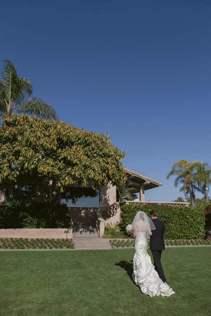 A bride in a white gown and veil walks arm-in-arm with a man in a black suit across a green lawn toward a house, with trees and a clear blue sky in the background.