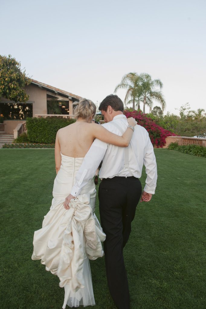A bride and groom walk arm-in-arm across a green lawn toward a house, with the bride holding up her dress. They are photographed from behind on a sunny day.