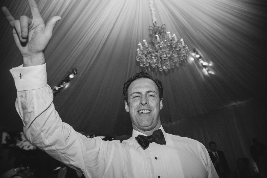 A man in a tuxedo smiles and raises his hand in a rock and roll gesture under a draped ceiling with a chandelier, at what appears to be a formal event or celebration.