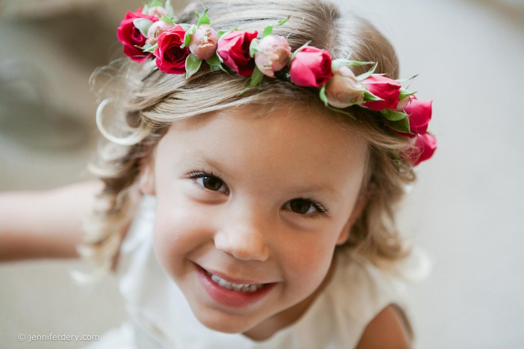 A young child with blonde hair smiles up at the camera, wearing a white dress and a floral crown made of red and pink roses. The background is softly blurred.