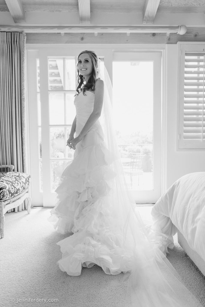 A bride in a ruffled wedding dress stands and smiles in a bright, sunlit room with large windows and French doors behind her. The room has a bed, a patterned bench, and light streaming in.