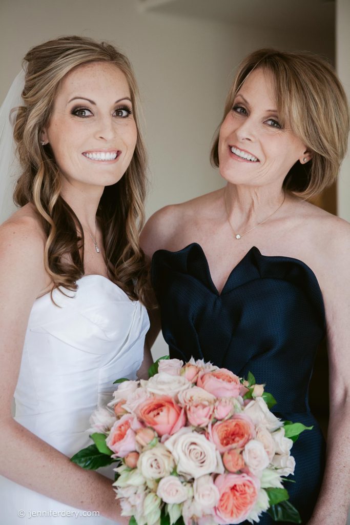 A bride in a white dress stands smiling next to a woman in a strapless navy dress, holding a bouquet of pink and peach roses. Both women are smiling and looking at the camera.
