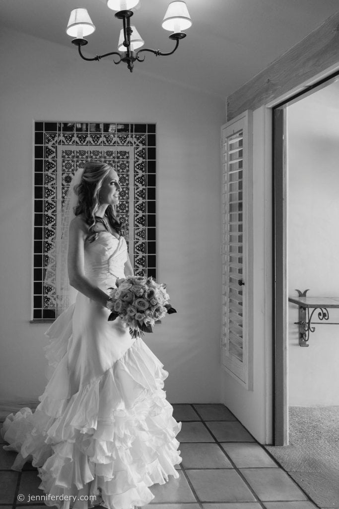 A bride in a ruffled wedding gown holds a bouquet and gazes out a window, standing by a tiled wall under a chandelier in a softly lit room.