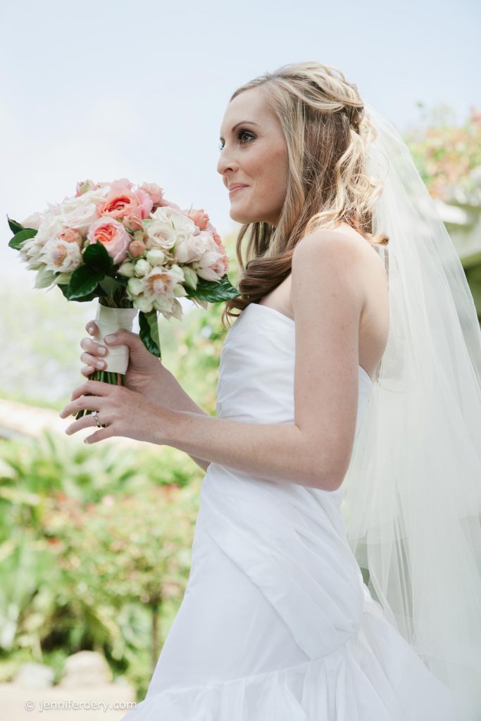 A bride in a white dress and veil holds a bouquet of pink and white flowers, standing outdoors with greenery in the background.