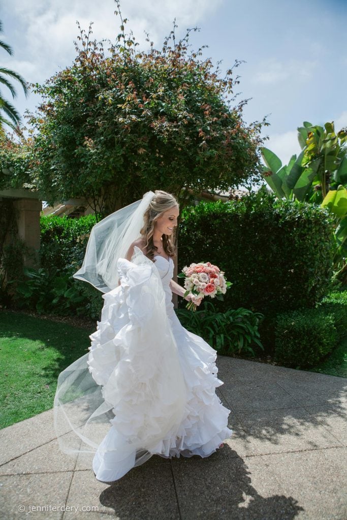 A bride in a white wedding dress and veil walks outdoors on a sunny day, holding a bouquet of pink flowers and lifting her dress slightly as she moves along a garden path.