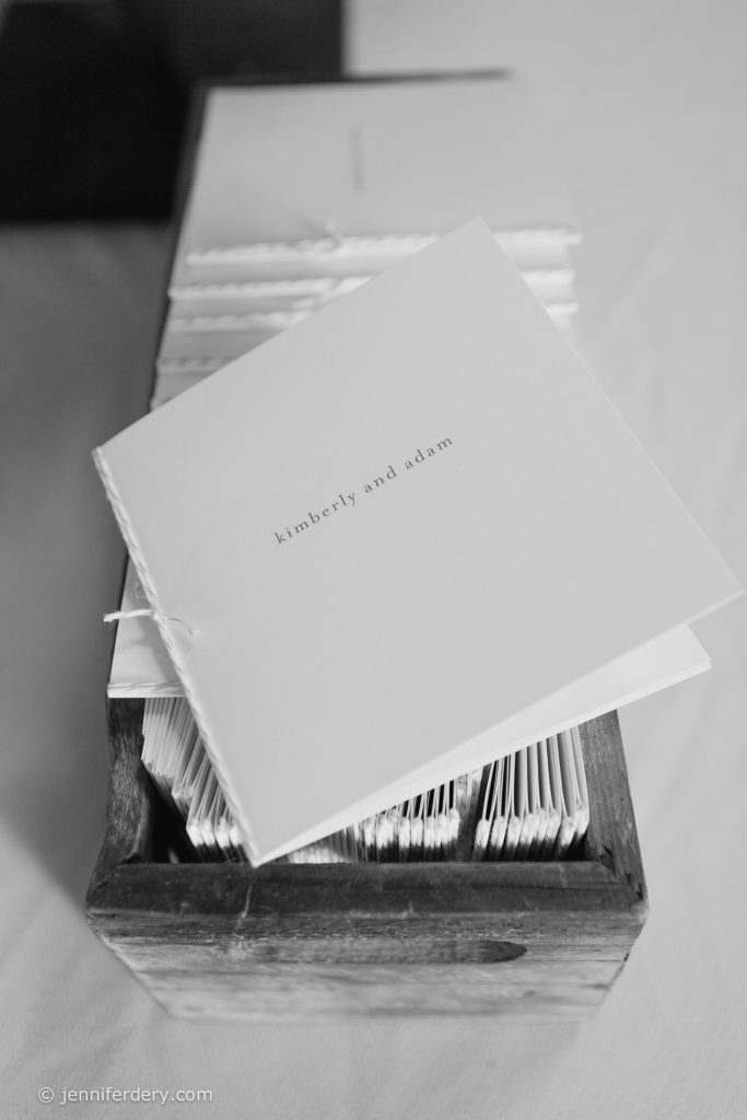 A stack of white booklets with “kimberly and adam” printed on the cover, neatly arranged in a wooden box on a table.