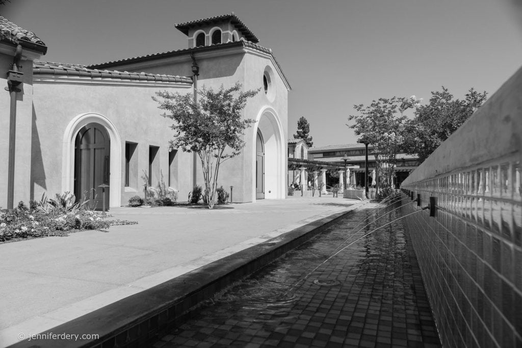 A black-and-white photo of a Mediterranean-style building with arched windows and a tiled roof. A narrow water feature runs along a tiled pathway, with streams of water arching into it. Trees and plants are visible in the background.