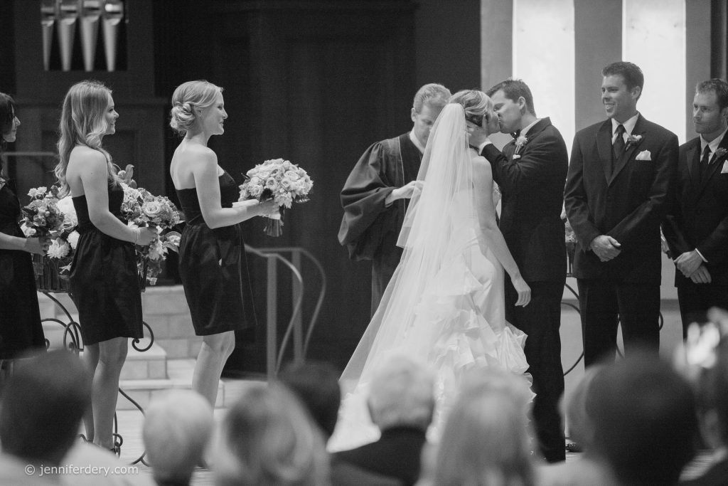 A bride and groom kiss at the altar during their wedding ceremony, surrounded by bridesmaids holding bouquets and groomsmen in suits, with an officiant standing behind them in a church.