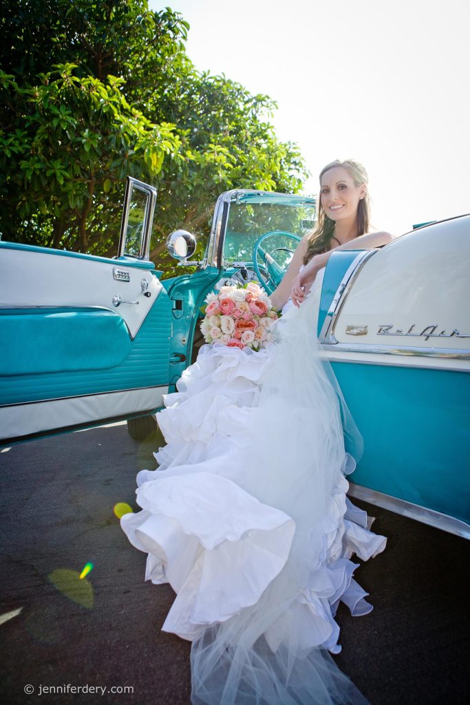 A bride in a white wedding dress sits in a vintage turquoise Chevrolet Bel Air, holding a bouquet of flowers, with the car door open and sunlight streaming through nearby trees.