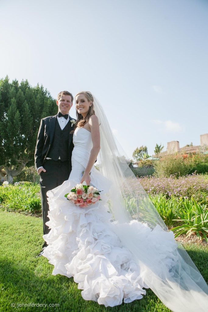 A bride in a white gown and veil stands next to a groom in a black tuxedo, smiling outdoors on a sunny day at Rancho Valencia Resort. The bride holds a bouquet of pink and white flowers, and they are surrounded by greenery and flowers.