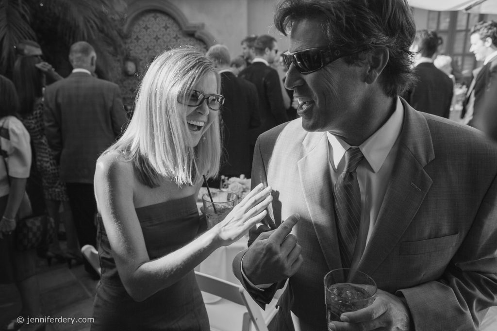 A woman and a man, both wearing sunglasses and formal attire, laugh and talk closely at a lively social gathering. The man holds a drink, and several people mingle in the background.