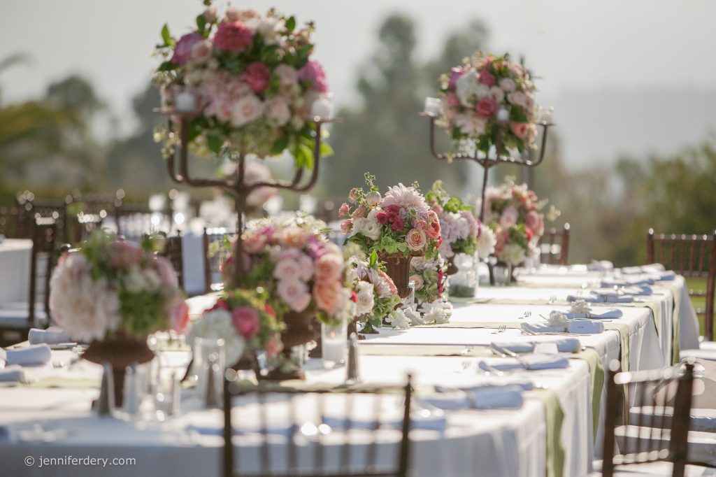 Elegant outdoor wedding reception setup with long tables, white tablecloths, and tall floral centerpieces featuring pink and white flowers. Brown chairs surround the tables under soft natural light.