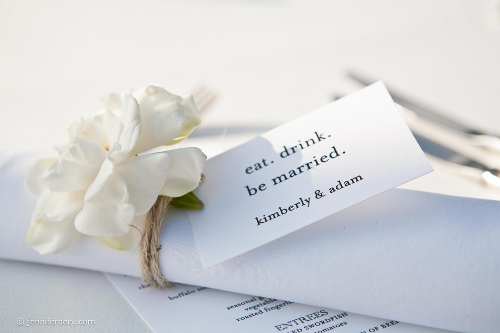 A white napkin with a white flower and twine is placed beside a card reading "eat. drink. be married. kimberly & adam." The setting appears elegant and is likely for a wedding celebration.