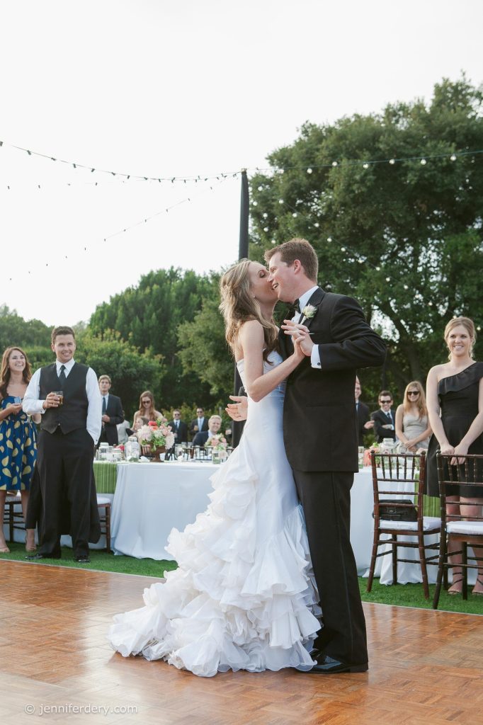 A bride and groom share a kiss on a wooden dance floor outdoors, surrounded by guests in formal attire. The bride wears a flowing white dress, and the groom is in a black tuxedo. Trees and string lights are in the background.