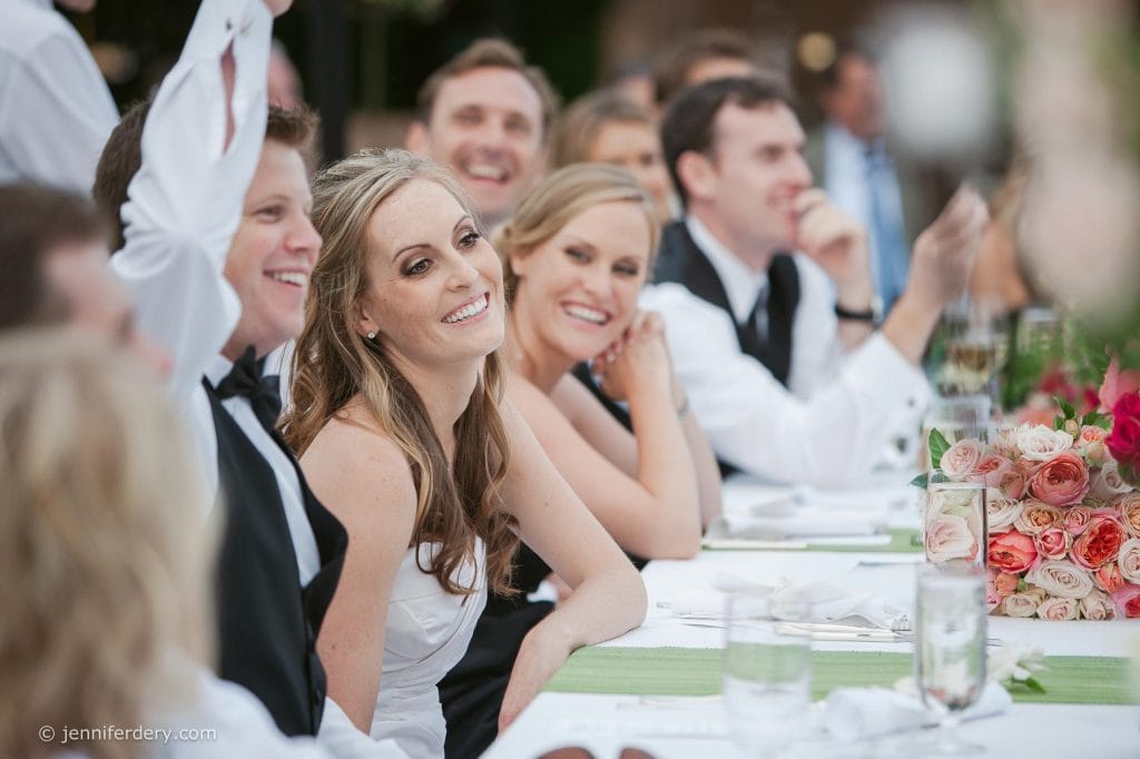 A joyful bride in a white dress smiles, seated at a long table with guests in formal attire. The group appears to be at an outdoor wedding reception, surrounded by flowers and elegant table settings.