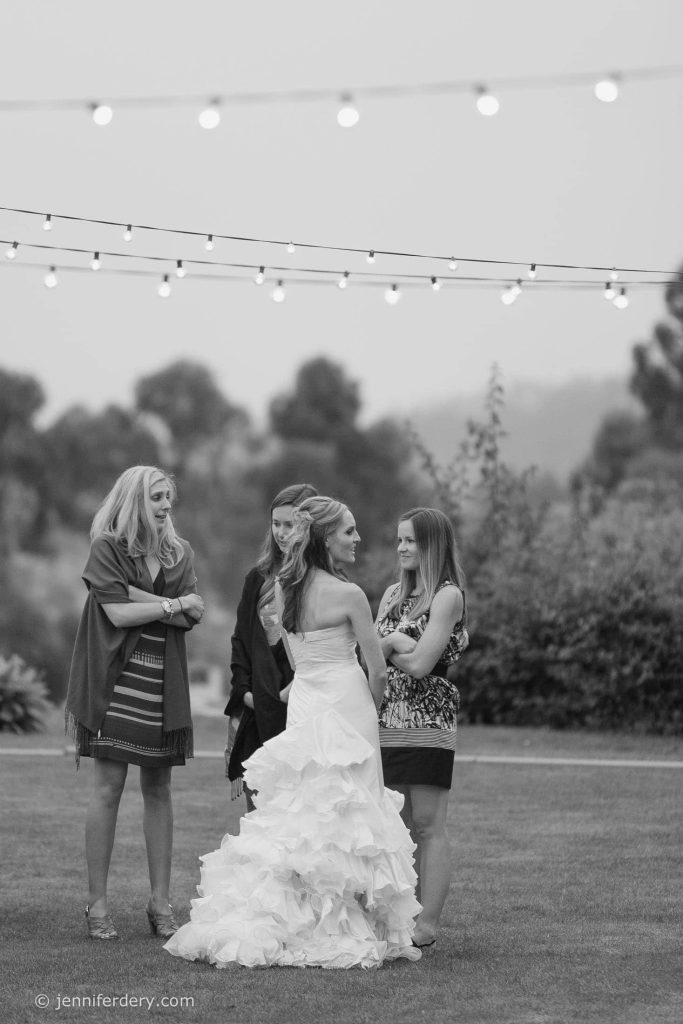 A bride in a white gown stands outdoors on grass, speaking with three women under string lights. The background is blurred with greenery and trees. The mood appears relaxed and conversational.