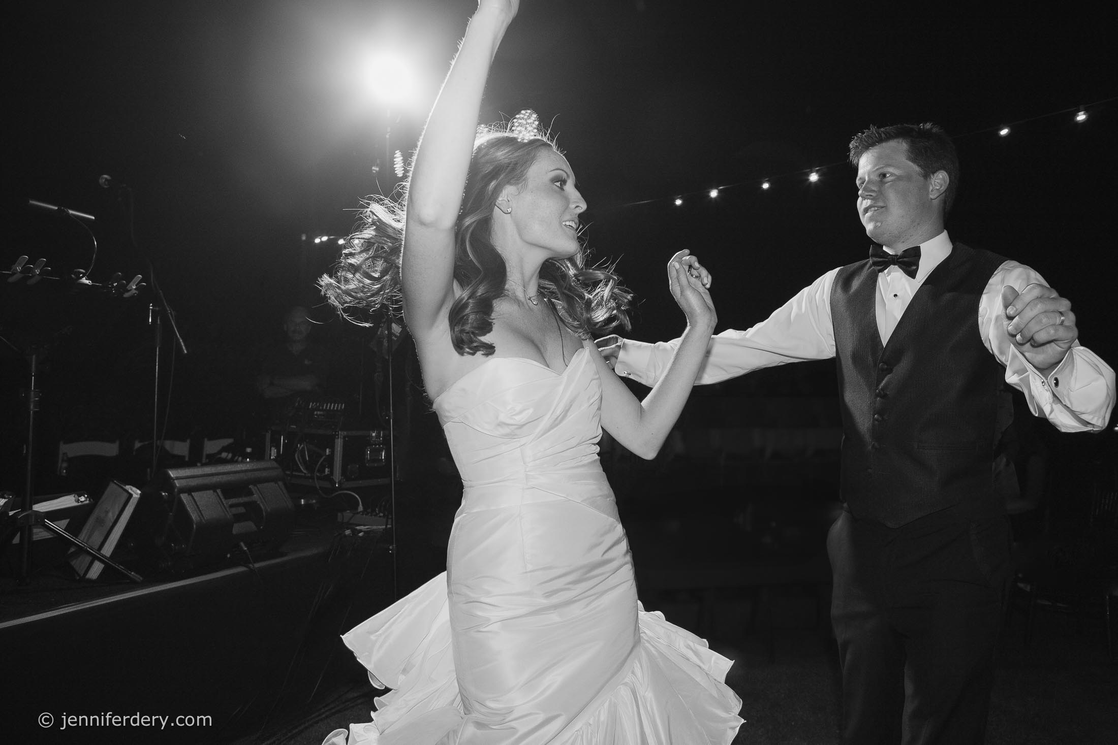 A woman in a white gown and a man in a tuxedo dance energetically at night under string lights, captured in mid-motion with the woman's hair and dress swirling.