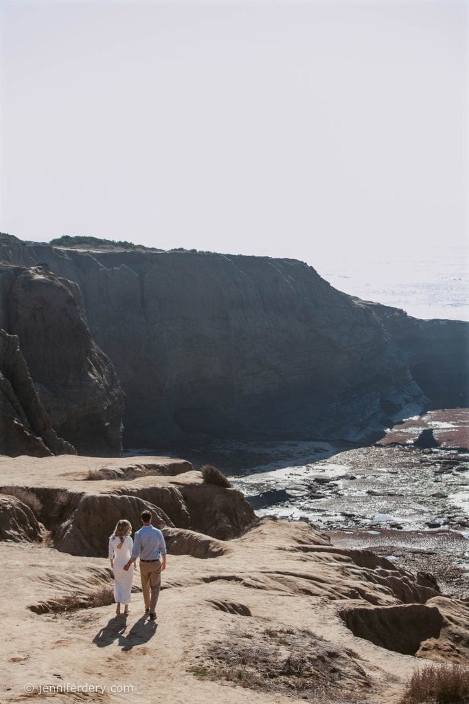 A couple walks hand in hand on a rocky, sunlit cliff overlooking the ocean, with large dark cliffs and the sea in the background.