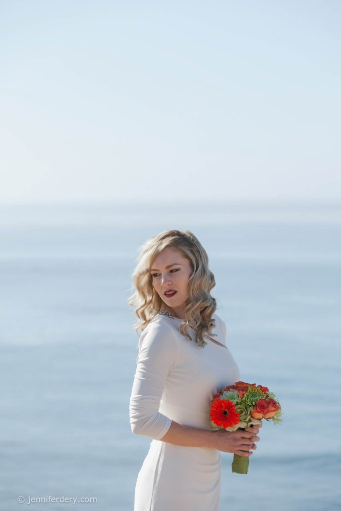 A woman in a white dress stands by the ocean, holding a bouquet of bright orange and red flowers and looking over her shoulder. The background is calm and blue with a soft, clear sky.